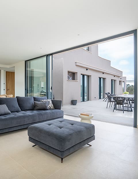 A modern living room with a dark grey sofa, light stone flooring, and sliding glass walls opening to an outdoor terrace.