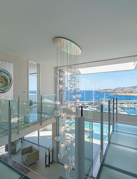 A glass-floored landing overlooks a double-height living space with a large feature chandelier and views of a marina.