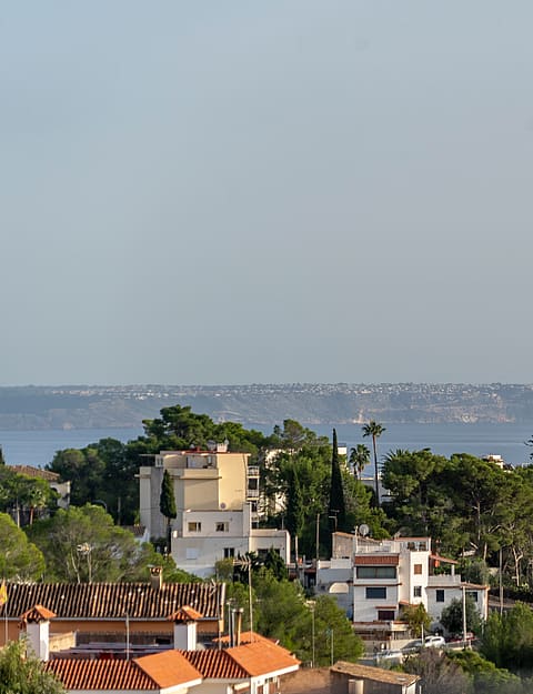 A wide landscape view looking out over trees and Mediterranean style houses towards a distant coastline and sea.