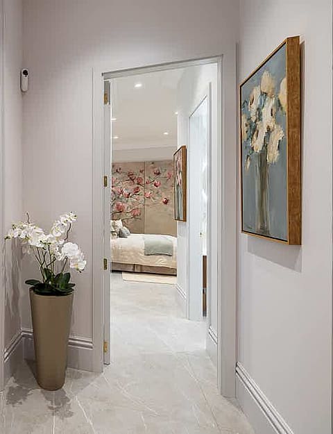 A bright hallway with white walls and stone flooring connecting a bedroom to a living area, featuring a tall plant pot.