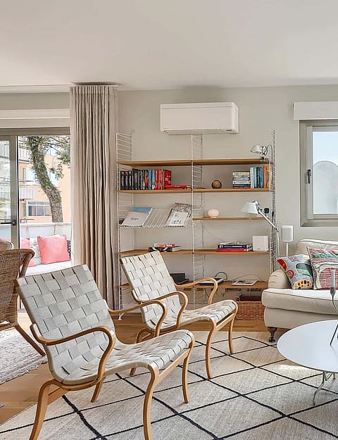Bright living room featuring a neutral sofa, patterned armchairs, metal shelving, and a large window leading to a terrace.