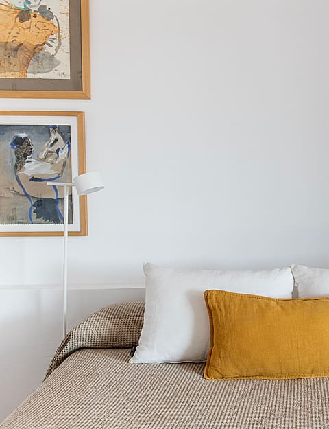 A double bed with a patterned beige cover and mustard yellow cushion, flanked by two minimalist floor lamps and framed wall art.