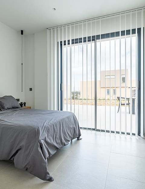 Modern bedroom with a grey bed, concrete floor, and vertical blinds covering a glass door to a terrace.