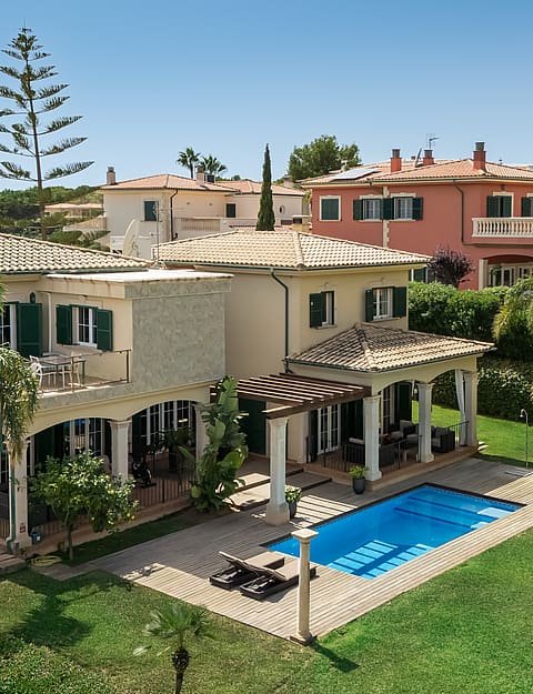A two-story villa with Mediterranean architecture, a tiled roof, green louvered shutters, and a swimming pool in the garden.