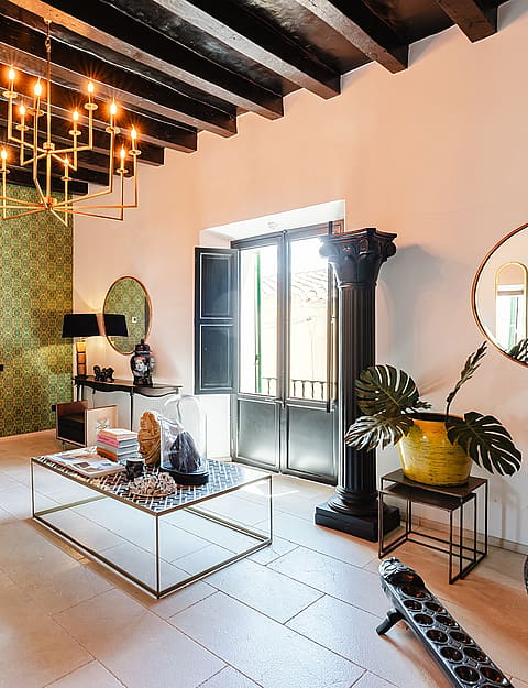 A spacious living room featuring a dark wooden beamed ceiling, a patterned green accent wall, and a large metal chandelier.