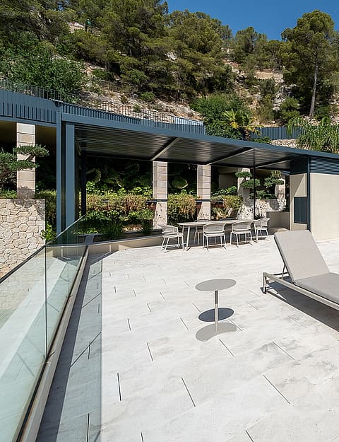 A wide outdoor terrace with stone paving, two sun loungers, and a covered dining area set against a hillside backdrop.