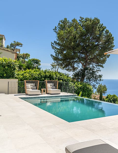 A rectangular swimming pool on a pale stone terrace overlooks the blue sea, framed by a pine tree and lounge furniture.