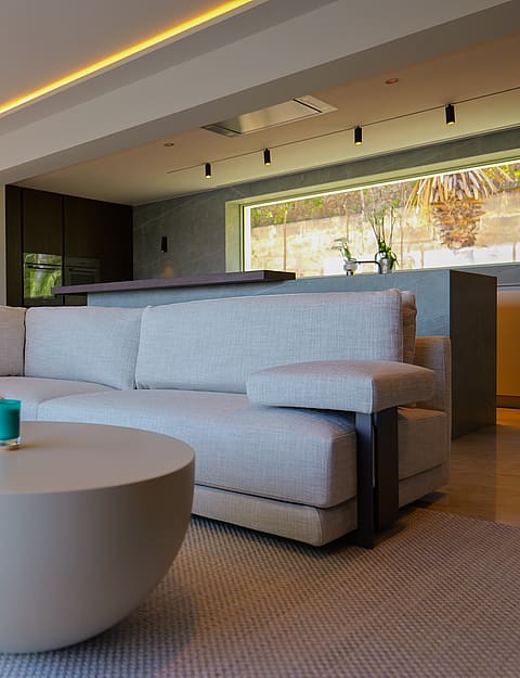 A contemporary open-plan living room featuring a light grey modular sofa, a rounded coffee table, and an integrated kitchen with a dark stone island and horizontal window.