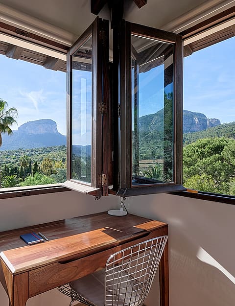 A small wooden desk positioned in a corner with floor-to-ceiling windows framing a landscape of Alaro mountains.