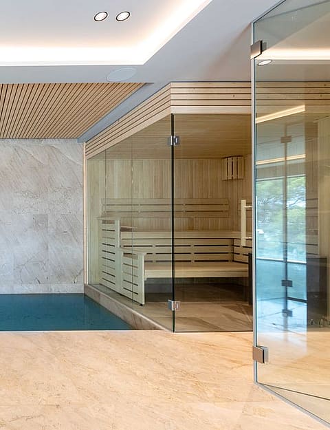 Indoor pool and sauna area with light wood slatted ceiling, large stone floor tiles, and glass partitions.