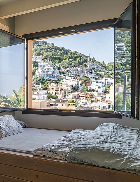 Interior bedroom view featuring an open casement window overlooking a hillside town in Genova with mountain slopes visible.