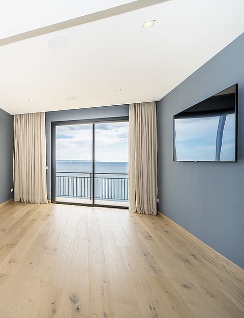 An empty bedroom with blue walls, light oak wood floors, and large glass sliding doors opening to a balcony with sea views.