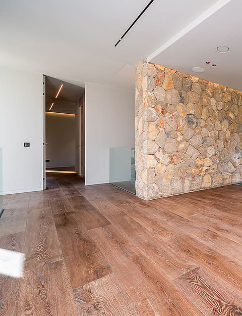Empty modern living area with light oak floors and a large dry-stone accent wall facing floor-to-ceiling windows.