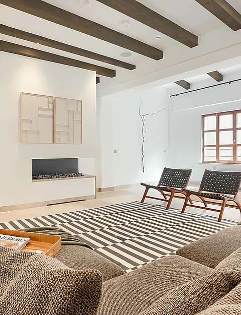 A living room interior featuring dark wood exposed ceiling beams, a textured stone wall, a large flat-screen TV on a wooden tripod stand, and two mid-century style armchairs on a patterned rug.
