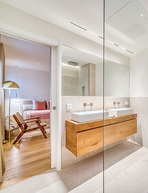 Interior view of a contemporary bathroom featuring a floating light oak vanity, dual ceramic basins, and a glass-enclosed shower with textured stone tiles.