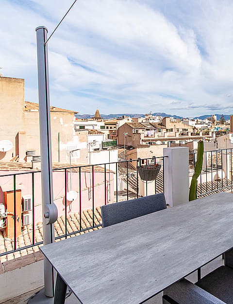 An outdoor terrace in an urban setting featuring a dining table, grey upholstered seating, and panoramic views of old town rooftops under a blue sky.