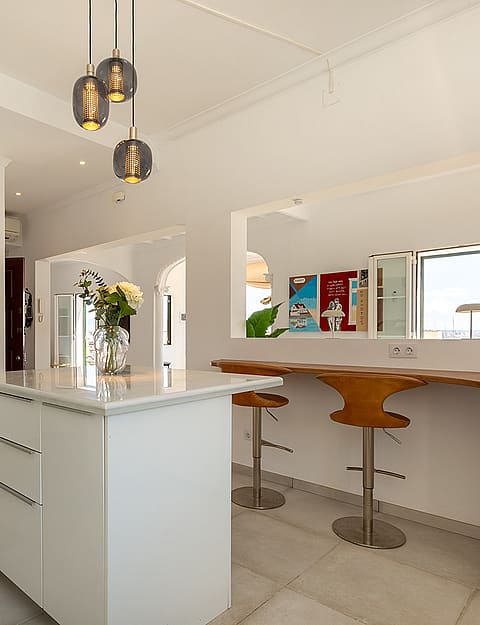A bright kitchen area with a white marble-topped island, wooden bar stools, and modern pendant lighting in a minimalist space.