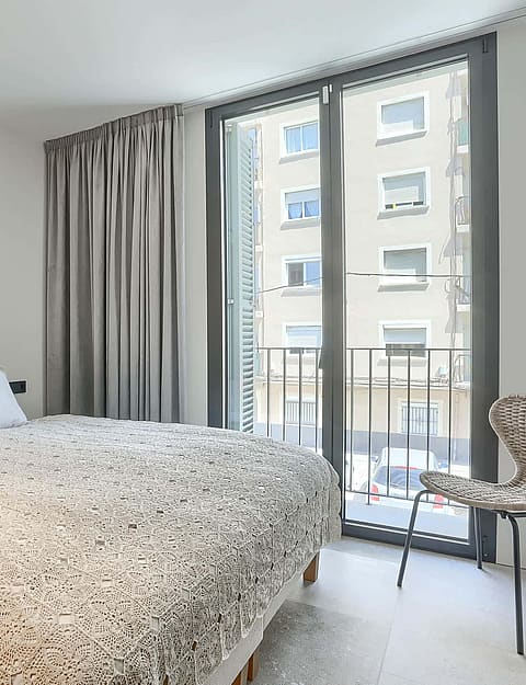 A contemporary bedroom featuring a double bed, patterned bedding, grey curtains, and a Juliet balcony overlooking a street.