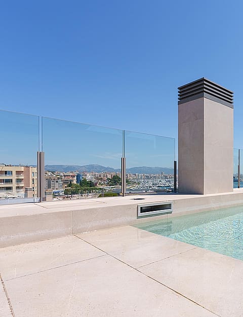 Piscina en azotea con barandillas de cristal hacia el mar, con pavimento de piedra y chimenea central.