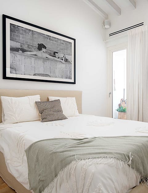 A bright bedroom with a queen-sized bed, white linens, a neutral headboard, framed black and white art, and a view into an en-suite bathroom.