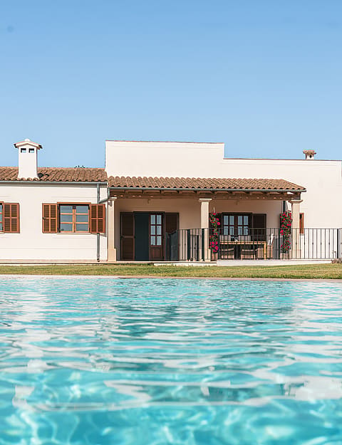 Vista de una finca blanca de una planta con tejado de teja árabe junto a una piscina azul bajo el cielo despejado.
