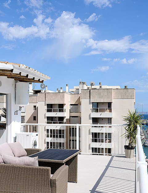 A rooftop terrace with rattan seating, overlooking a marina with boats and nearby apartment buildings under a blue sky.