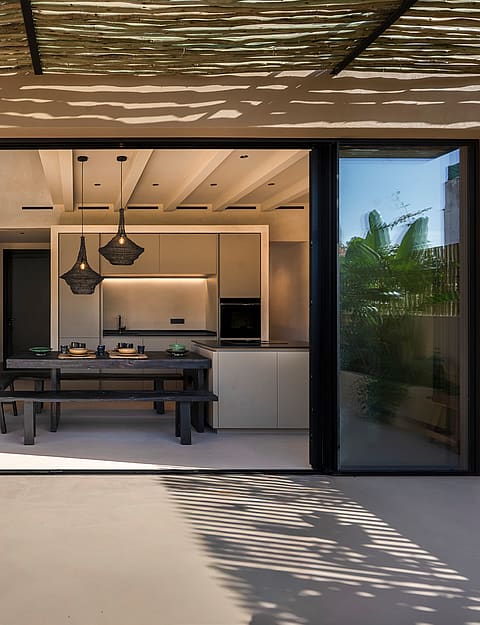 A modern kitchen and dining space viewed from a terrace through large sliding glass doors, featuring plastered walls and wooden ceiling beams.
