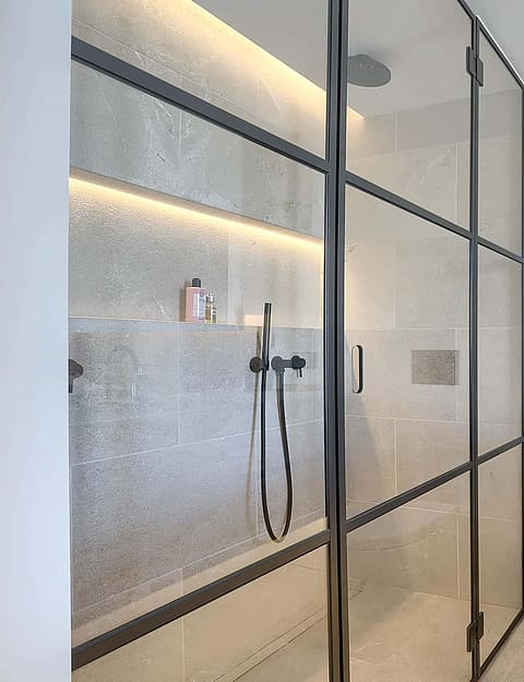A bathroom featuring a walk-in shower with a black metal-framed glass partition, a stone-composite sink, and minimalist white walls.