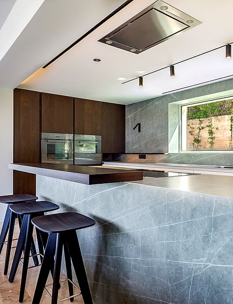 Modern kitchen with a large grey marble island, wooden cabinets, and three dark wooden bar stools under track lighting.