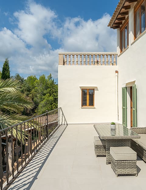 An elevated Mediterranean terrace featuring wicker furniture, wrought iron railings, and views of lush green trees.