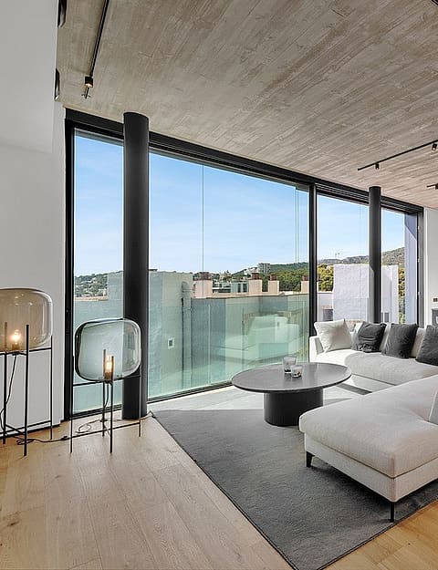 A modern open-plan living room with a grey sectional sofa, oak wood floors, and a textured concrete ceiling, leading to a glass-railed terrace with sea views.
