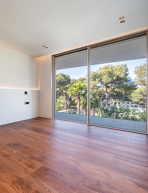 Empty bedroom with white walls, oak flooring, and sliding glass doors opening to a lush garden view.