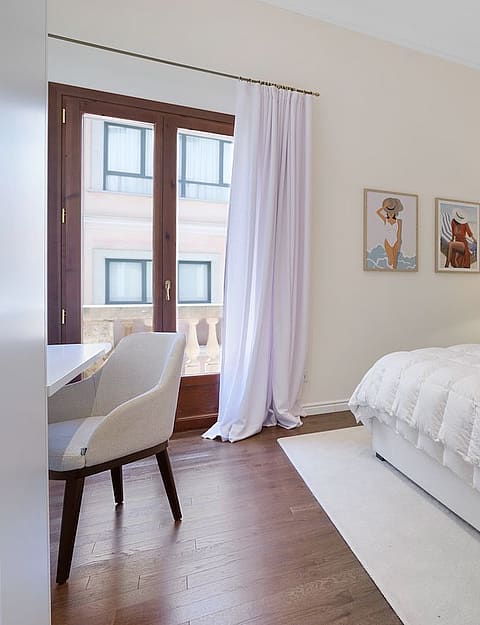 A bright bedroom with a king-sized bed, white linens, patterned wallpaper behind the headboard, and a balcony with white curtains.