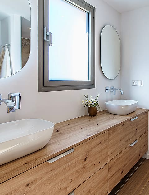 Modern bathroom featuring a long wooden vanity with two white ceramic vessel sinks, circular mirrors, and wooden flooring.