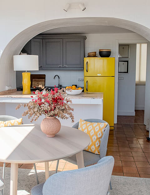 A small kitchen area with a yellow refrigerator, dark wooden cabinets, and a terracotta tiled floor seen through a wide arch.