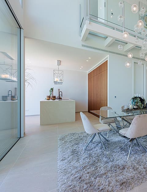 Dining room with a glass table, upholstered chairs on a textured rug, and a modern glass elevator in a double-height space.