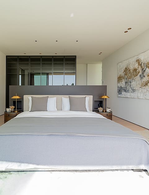 A minimalist master bedroom featuring a large bed, gray headboard, and abstract wall art, with an adjacent bathroom visible.