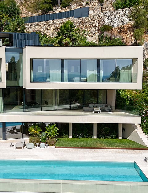 Exterior view of a multi-level contemporary villa built into a rocky hillside, featuring floor-to-ceiling glass walls, a rectangular swimming pool, and terraced gardens.