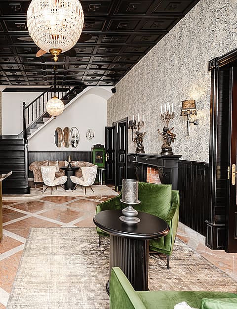 A spacious, eclectic interior featuring a large bar with metallic front, patterned wallpaper, black coffered ceiling, and a central staircase leading to the upper level.
