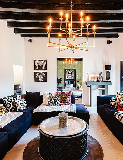 Interior view of a living room with dark low-profile sofas, a central round metal coffee table, and an ornate brass chandelier hanging from dark exposed ceiling beams.