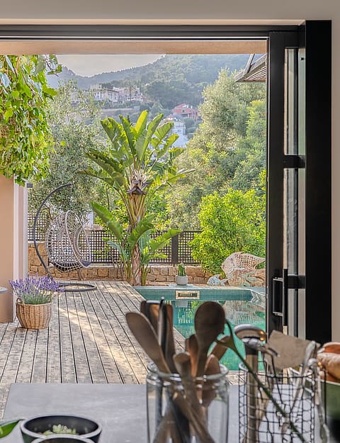 Interior view from a kitchen counter looking out through an open glass door to a stone-paved terrace with lush greenery.