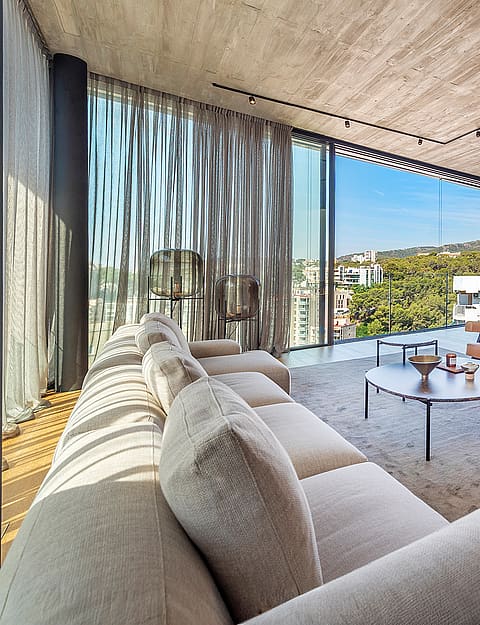 Wide-angle shot of a bright, open-plan living room with exposed concrete ceilings, light wood floors, and floor-to-ceiling glass walls looking out to coastal scenery.