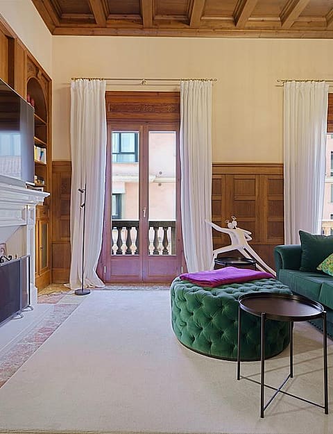 A formal living area featuring dark wood wall paneling, a stone fireplace, a green velvet sofa, and tall windows facing the city.