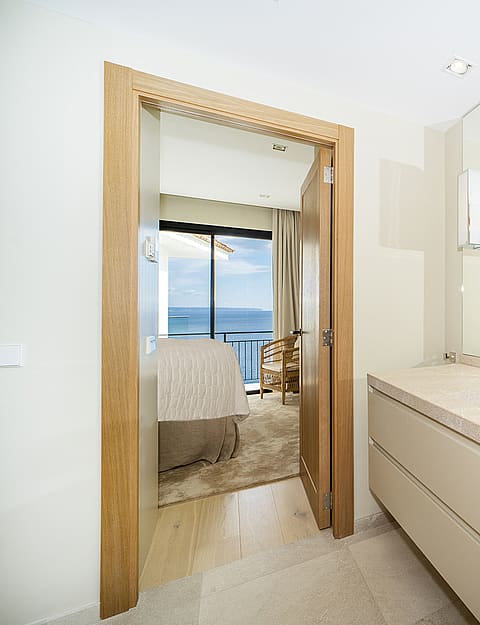 A modern bathroom vanity with a light stone countertop, a large frameless mirror, and a view through an open doorway into a bedroom overlooking the sea.