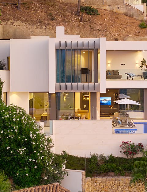 Exterior aerial view of a multi-level contemporary villa built into a rocky hillside with tiered terraces, pool, and lighting.