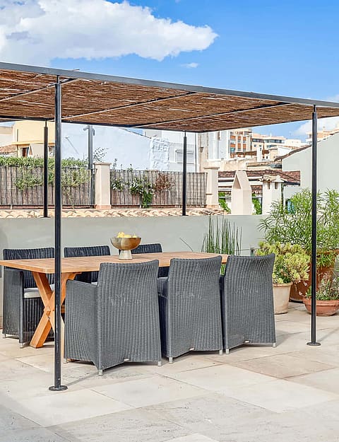 Rooftop terrace with a wooden dining table, woven chairs, and a metal pergola, surrounded by potted succulents and city views.