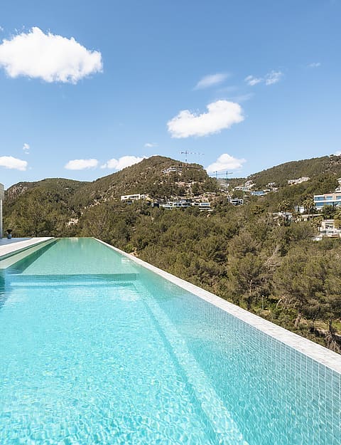 A long turquoise infinity pool runs alongside a modern villa with glass walls, overlooking a verdant mountain range.