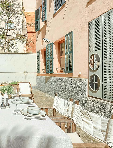 An outdoor dining table set with white linens beside a terracotta wall with green shutters, surrounded by palm trees.