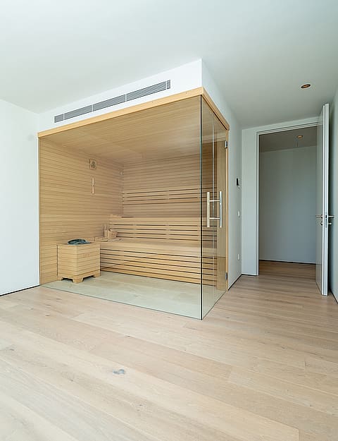 Modern home sauna with light wood paneling and glass doors inside a minimalist white room with light oak flooring.