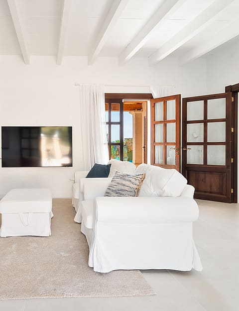 Salón con sofás blancos, otomana central y vigas de techo pintadas de blanco.
